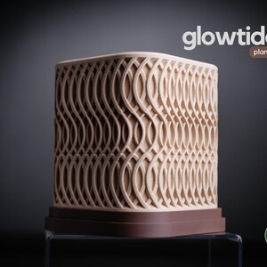 May include: A beige planter with a repeating wave-like pattern. The planter has a square shape and sits on a dark brown base. The word "glowtide" is above the planter, with "planter" below it.