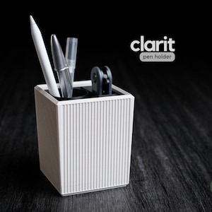 May include: A white, ribbed pen holder with the text "clarit pen holder" on a black background. The pen holder contains a white pen, a silver pen, and a clear pen. The pen holder is a cube shape.
