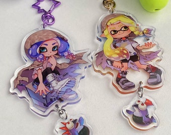 Könnte beinhalten: Zwei bunte Acryl-Schlüsselanhänger mit Comicfiguren aus dem Videospiel Splatoon. Ein Schlüsselanhänger zeigt eine lila und blaue Figur mit einem braunen Kapuzenmantel und einem blau-violetten Oktopus. Der andere Schlüsselanhänger zeigt eine gelbe Figur mit einem weißen Kapuzenmantel und einem gelben Oktopus. Beide Schlüsselanhänger haben einen kleinen Anhänger, der an der Unterseite befestigt ist.