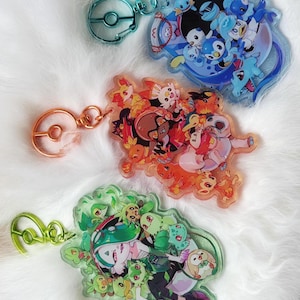 May include: Three colorful keychains featuring different Pokemon characters. Each keychain has a metal ring with a Pokeball design. The first keychain features a blue and white character with a group of blue and white Pokemon. The second keychain features a red and orange character with a group of orange and red Pokemon. The third keychain features a green and white character with a group of green and white Pokemon.