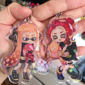 May include: Two iridescent acrylic keychains featuring anime-style characters from the video game Splatoon. The keychain on the left features a girl with orange hair and a black and white squid. The keychain on the right features a girl with red hair and a pink squid.