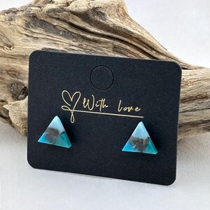 Op de afbeelding: Een paar turquoise en zwarte driehoekige oorstekers op een zwarte kaart. De kaart heeft de woorden "With love" in gouden letters. De oorbellen worden gepresenteerd tegen een achtergrond van drijfhout.