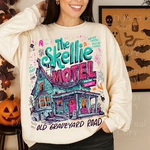 K&ouml;nnte beinhalten: Cremefarbenes Sweatshirt mit einem gruseligen Motel-Motiv. Der t&uuml;rkisfarbene und rosa Aufdruck lautet "The Skellie Motel" mit einer Illustration eines Spukhauses. Die Worte "Old Graveyard Road" stehen unten.