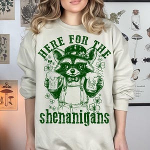 Puede incluir: Una sudadera de color beige claro con un gr&aacute;fico verde de un mapache sosteniendo dos jarras de cerveza. El texto "Here for the shenanigans" est&aacute; impreso debajo del mapache.