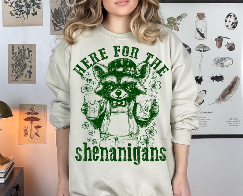 Puede incluir: Una sudadera de color beige claro con un gr&aacute;fico verde de un mapache sosteniendo dos jarras de cerveza. El texto "Here for the shenanigans" est&aacute; impreso debajo del mapache.