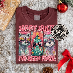 Puede incluir: Camiseta burdeos con el texto "SORRY SANTA I'VE BEEN FERAL" y un gr&aacute;fico de tres mapaches con gafas de sol y un &aacute;rbol de Navidad. Incluye un sello de "DO NOT COPY".