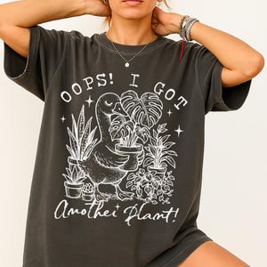 May include: A dark grey t-shirt with a white graphic of a goose holding a potted plant. The text "Oops! I got another plant!" is printed on the shirt.