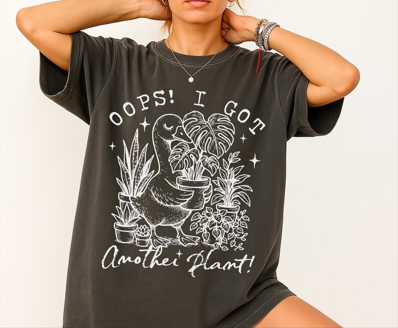 May include: A dark grey t-shirt with a white graphic of a goose holding a potted plant. The text "Oops! I got another plant!" is printed on the shirt.