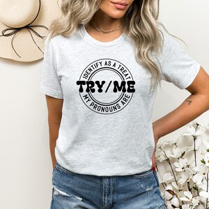 Puede incluir: Camiseta gris con un gr&aacute;fico circular negro que dice "Identify as a treat, Try/Me, My pronouns are".
