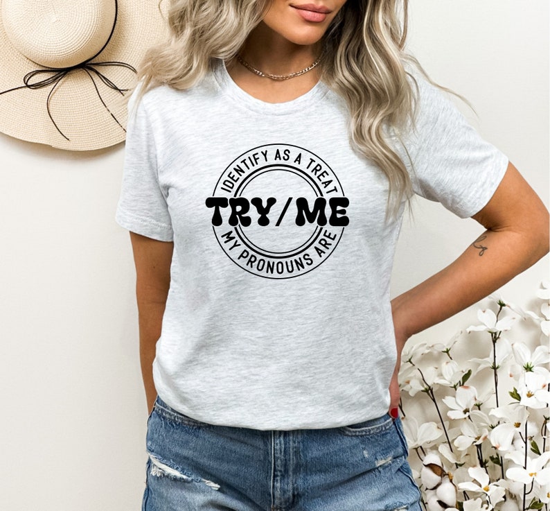Puede incluir: Camiseta gris con un gr&aacute;fico circular negro que dice "Identify as a treat, Try/Me, My pronouns are".