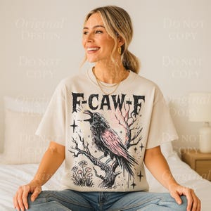 May include: Cream-colored t-shirt featuring a black crow perched on a branch, with pink accents and the text "F-CAW-F". The graphic design includes stars and flowers. The shirt is a casual, short-sleeved style.