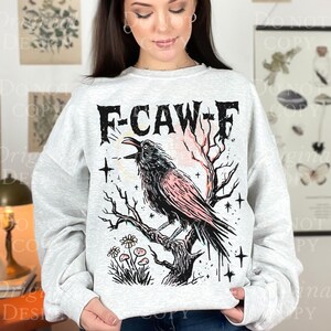May include: A light gray sweatshirt featuring a black and pink crow perched on a branch, with the text "F-CAW-F" above. The design includes stars, flowers, and a stylized sunrise. The sweatshirt is a comfortable, casual style.