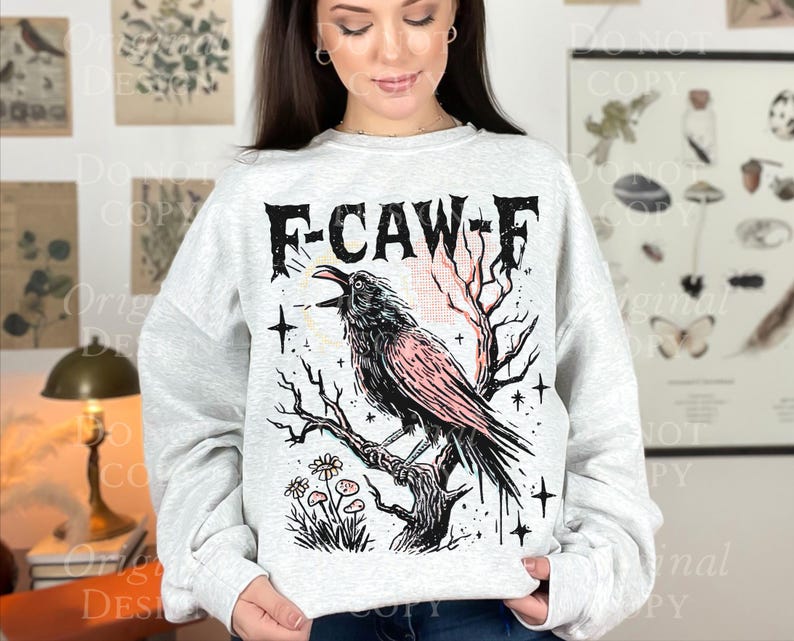 May include: A light gray sweatshirt featuring a black and pink crow perched on a branch, with the text "F-CAW-F" above. The design includes stars, flowers, and a stylized sunrise. The sweatshirt is a comfortable, casual style.