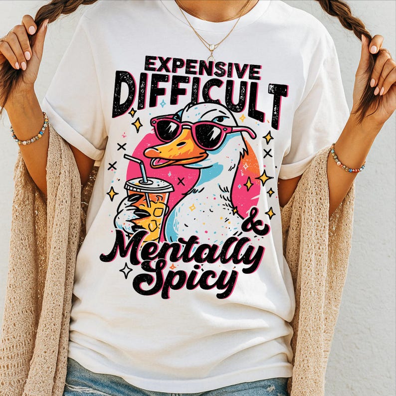 Pu&ograve; includere: T-shirt bianco con un'anatra dei cartoni animati con occhiali da sole e una bevanda. La maglietta ha la scritta "Expensive, Difficult & Mentally Spicy" in un carattere giocoso. L'anatra &egrave; ambientata su uno sfondo rosa e arancione con stelle.