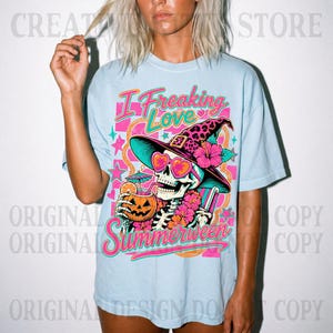 May include: A light blue t-shirt featuring a graphic of a skeleton wearing a leopard print witch hat and heart-shaped sunglasses. The skeleton holds a jack-o'-lantern drink with a straw. The text reads I Freaking Love Summerween.