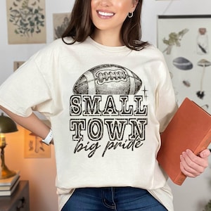 May include: A beige t-shirt with a vintage-style football graphic and the text "SMALL TOWN big pride".