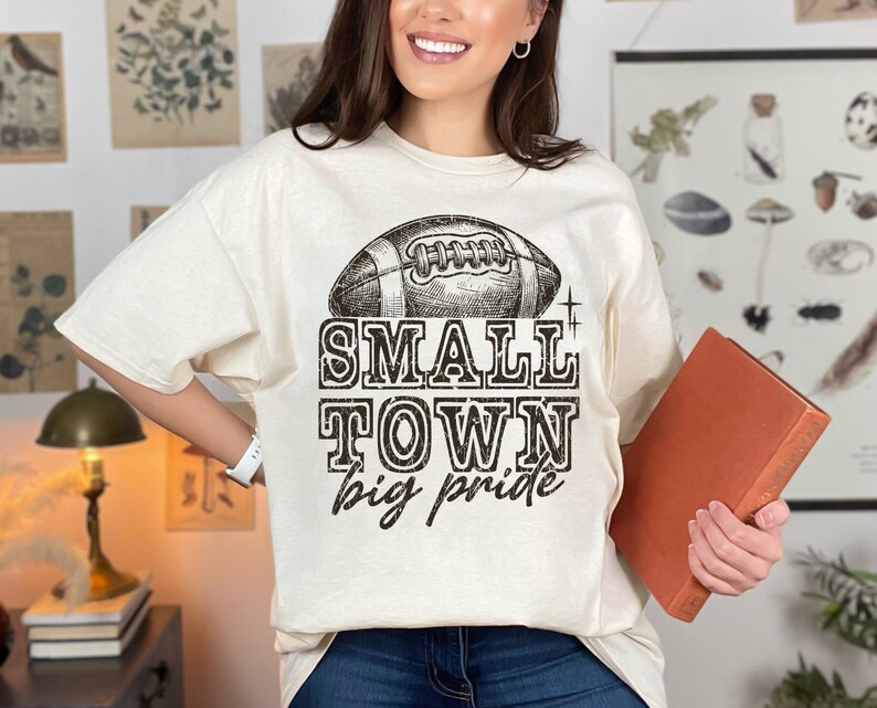 May include: A beige t-shirt with a vintage-style football graphic and the text "SMALL TOWN big pride".