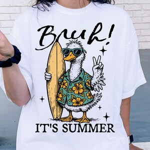 May include: White t-shirt with a cartoon duck wearing sunglasses and a floral shirt holding a surfboard. The text "Bruh! It's Summer" is printed on the shirt.