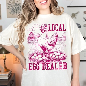 Op de afbeelding: Een wit T-shirt met een roze afbeelding van een kip met een mandje eieren en de tekst "LOCAL EGG DEALER".