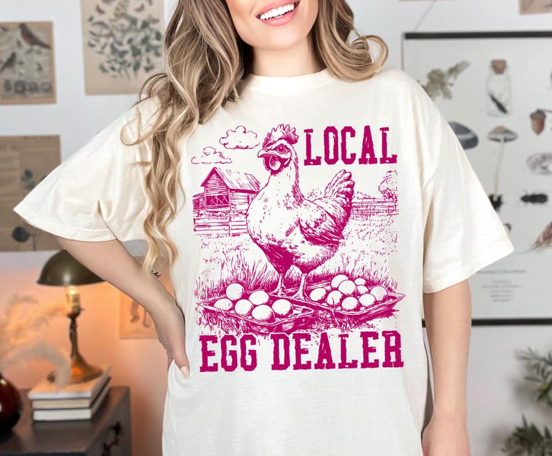 Op de afbeelding: Een wit T-shirt met een roze afbeelding van een kip met een mandje eieren en de tekst "LOCAL EGG DEALER".