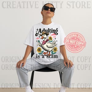 May include: White t-shirt featuring a cartoon duck in a cowboy hat and bandana, with sunflowers. The text "Adulting is a scam" is printed below the duck. A pink stamp reads "Original Design Do Not Copy!" in a circle.