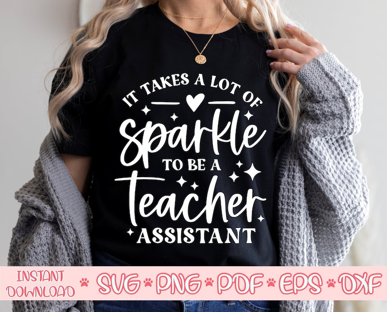 Op de afbeelding: Een zwart T-shirt met witte tekst die luidt "Het kost veel glitter om een ​​leraar-assistent te zijn". De tekst is omringd door kleine witte sterren en een roze hart.