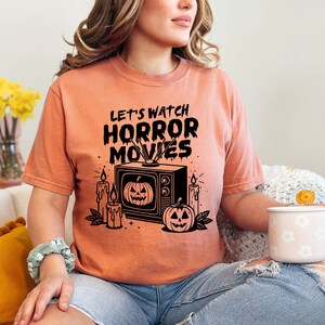 May include: A peach colored t-shirt with the text "Let's watch horror movies" in black. The graphic features a vintage television with a jack-o-lantern on the screen, candles, and a jack-o-lantern on the ground.