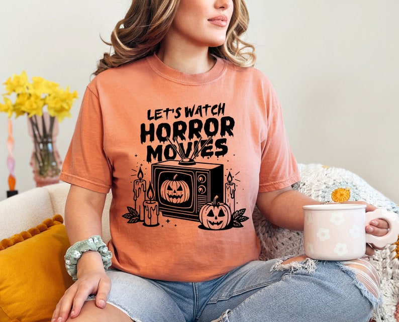 May include: A peach colored t-shirt with the text "Let's watch horror movies" in black. The graphic features a vintage television with a jack-o-lantern on the screen, candles, and a jack-o-lantern on the ground.