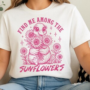 May include: White t-shirt with a pink graphic of a duckling holding a bunch of sunflowers. The text "Find me among the sunflowers" is printed in pink.