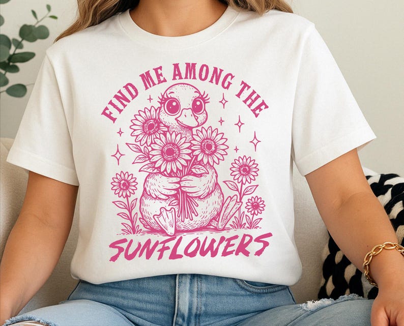 May include: White t-shirt with a pink graphic of a duckling holding a bunch of sunflowers. The text "Find me among the sunflowers" is printed in pink.