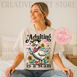 May include: A cream-coloured t-shirt featuring a duck in a cowboy hat holding a phone, with the text "Adulting is a scam". The design includes sunflowers and a pink cloud. The shirt has the text "Creative Kitty Store".