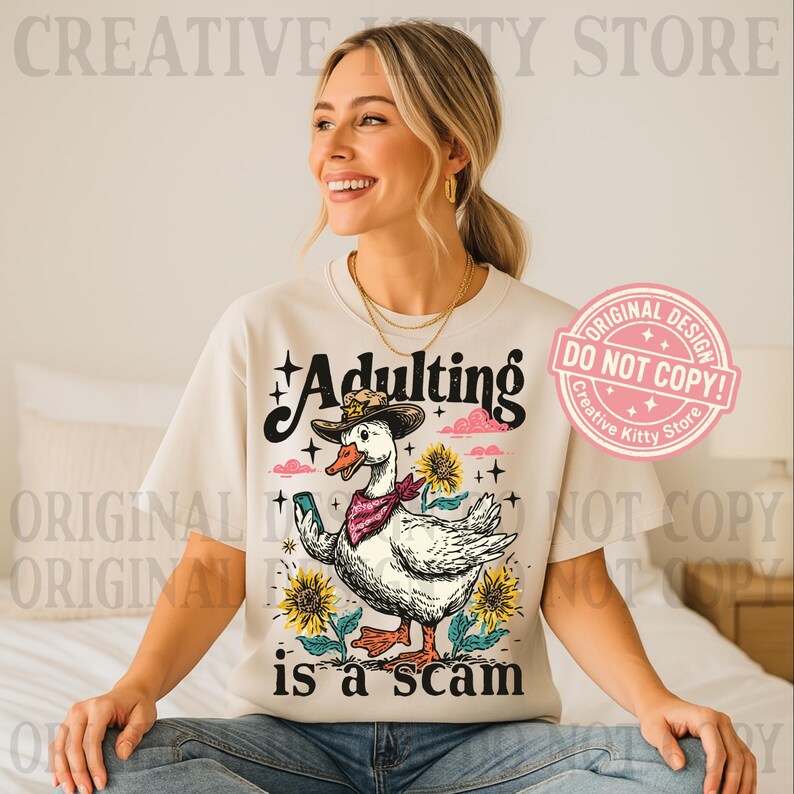 May include: A cream-coloured t-shirt featuring a duck in a cowboy hat holding a phone, with the text "Adulting is a scam". The design includes sunflowers and a pink cloud. The shirt has the text "Creative Kitty Store".