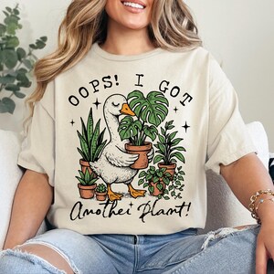 May include: A white t-shirt featuring a graphic of a goose holding a potted plant. The text on the shirt reads "Oops! I got another plant!"