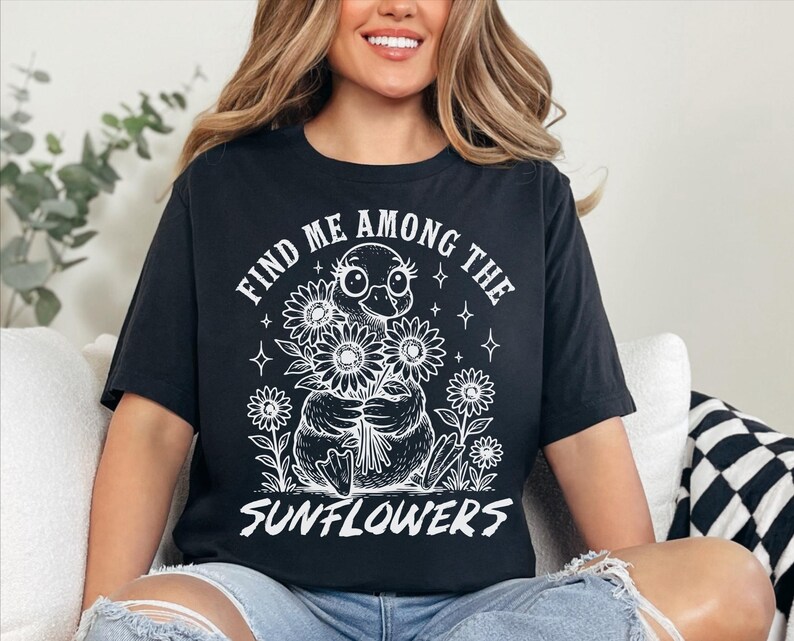 May include: Black t-shirt with a white graphic of a duck holding a bouquet of sunflowers. The text "Find me among the sunflowers" is printed on the shirt.