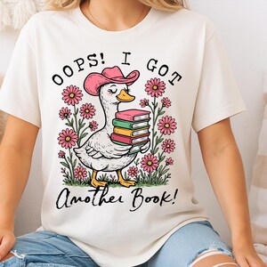May include: White t-shirt with a cartoon illustration of a duck wearing a pink cowboy hat and holding a stack of books. The text "Oops! I got another book!" is printed on the shirt.