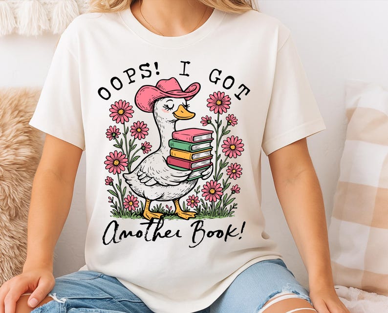 May include: White t-shirt with a cartoon illustration of a duck wearing a pink cowboy hat and holding a stack of books. The text "Oops! I got another book!" is printed on the shirt.