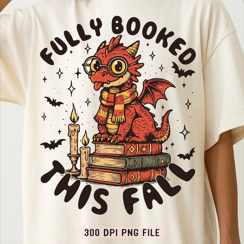 K&ouml;nnte beinhalten: Cremefarbenes T-Shirt mit einem Cartoon-Drachen mit Brille und Schal, der auf B&uuml;chern sitzt. Der Text "FULLY BOOKED THIS FALL" steht &uuml;ber und unter dem Drachen. Flederm&auml;use und Sterne sind ebenfalls im Design enthalten.