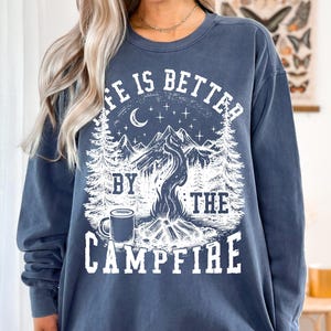 Puede incluir: Una sudadera azul marino con un gr&aacute;fico blanco que dice "LIFE IS BETTER BY THE CAMPFIRE". El dise&ntilde;o incluye una fogata, monta&ntilde;as, &aacute;rboles, una luna, estrellas y una taza. La sudadera es de corte holgado.