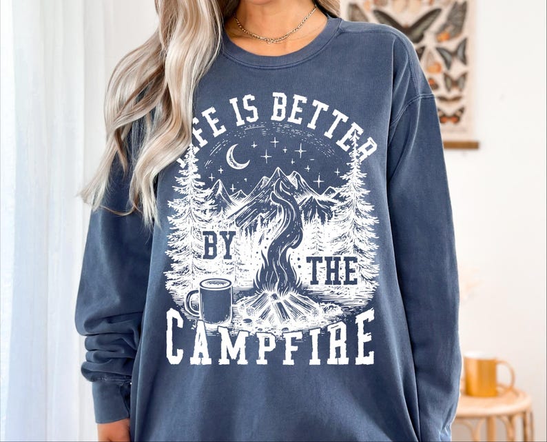 Puede incluir: Una sudadera azul marino con un gr&aacute;fico blanco que dice "LIFE IS BETTER BY THE CAMPFIRE". El dise&ntilde;o incluye una fogata, monta&ntilde;as, &aacute;rboles, una luna, estrellas y una taza. La sudadera es de corte holgado.