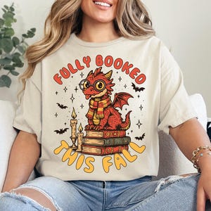 K&ouml;nnte beinhalten: Beiges T-Shirt mit einem roten Drachen mit Brille und Schal, der auf B&uuml;chern sitzt. Der Text "FULLY BOOKED THIS FALL" ist orange. Flederm&auml;use und Sterne umgeben den Drachen.