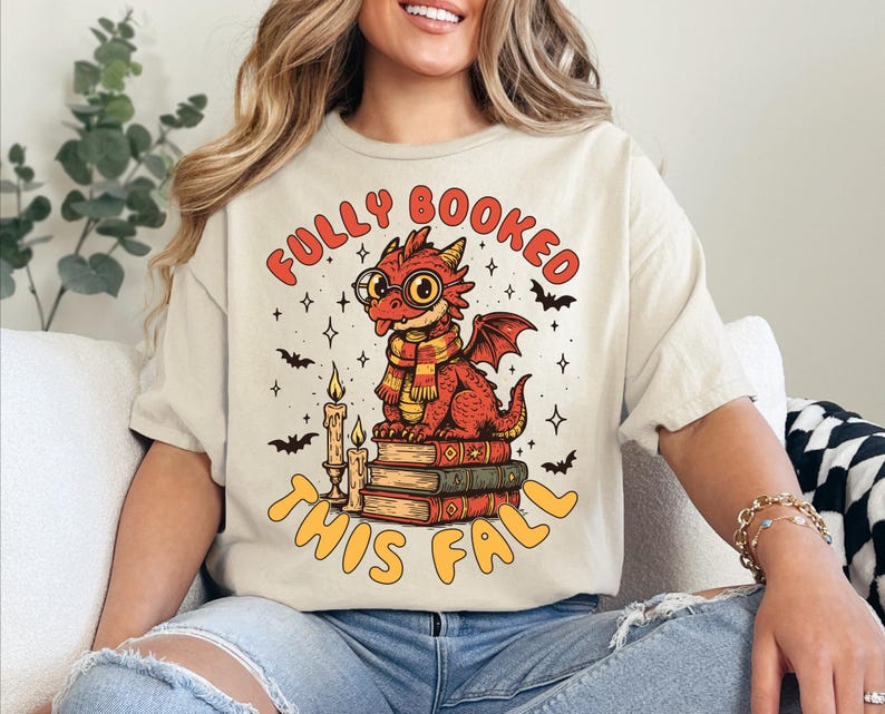 K&ouml;nnte beinhalten: Beiges T-Shirt mit einem roten Drachen mit Brille und Schal, der auf B&uuml;chern sitzt. Der Text "FULLY BOOKED THIS FALL" ist orange. Flederm&auml;use und Sterne umgeben den Drachen.