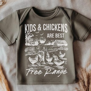 K&ouml;nnte beinhalten: Ein gr&uuml;nes T-Shirt mit kurzen &Auml;rmeln und einem wei&szlig;en Aufdruck eines Kindes und H&uuml;hnern. Der Aufdruck lautet "Kids & Chickens Are Best Free Range".