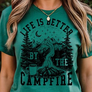 Puede incluir: Camiseta verde con un gr&aacute;fico negro de una escena de fogata. El dise&ntilde;o incluye monta&ntilde;as, &aacute;rboles, una luna creciente, estrellas y una taza de caf&eacute;. El texto "LIFE IS BETTER BY THE CAMPFIRE" est&aacute; impreso.