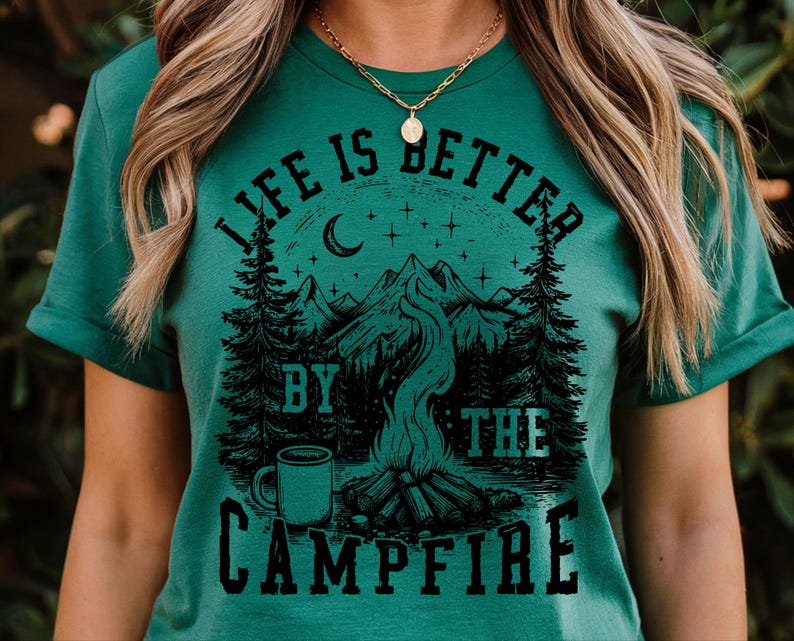 Puede incluir: Camiseta verde con un gr&aacute;fico negro de una escena de fogata. El dise&ntilde;o incluye monta&ntilde;as, &aacute;rboles, una luna creciente, estrellas y una taza de caf&eacute;. El texto "LIFE IS BETTER BY THE CAMPFIRE" est&aacute; impreso.