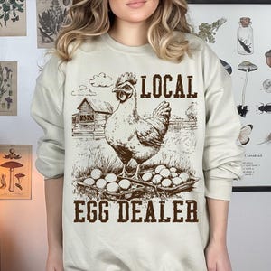 Op de afbeelding: Een beige sweatshirt met een bruine afbeelding van een kip en eieren. De tekst "LOCAL EGG DEALER" is op het sweatshirt gedrukt.