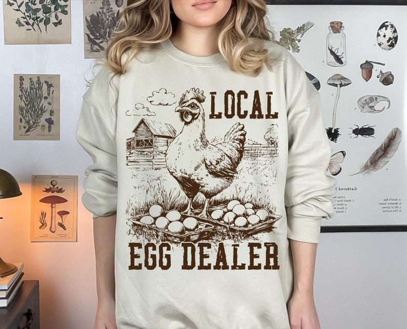 Op de afbeelding: Een beige sweatshirt met een bruine afbeelding van een kip en eieren. De tekst "LOCAL EGG DEALER" is op het sweatshirt gedrukt.