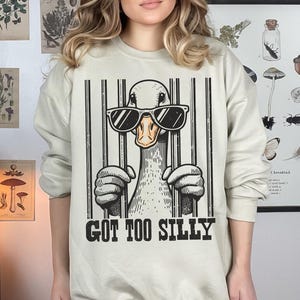 Pu&ograve; includere: Una felpa beige con una grafica di un'anatra che indossa occhiali da sole e dietro le sbarre. Il testo "GOT TOO SILLY" &egrave; stampato sotto la grafica.