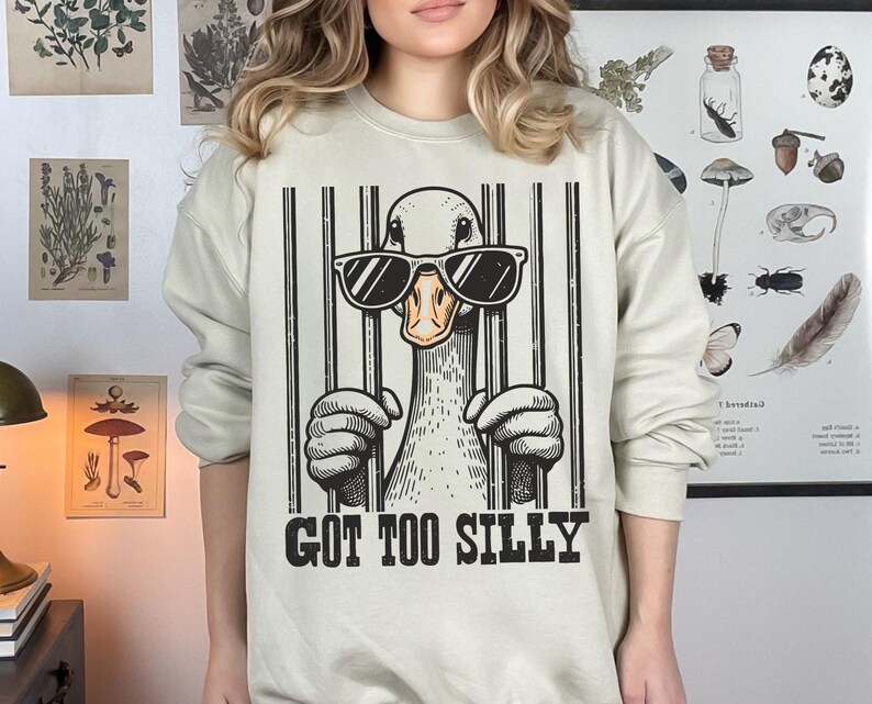 Pu&ograve; includere: Una felpa beige con una grafica di un'anatra che indossa occhiali da sole e dietro le sbarre. Il testo "GOT TOO SILLY" &egrave; stampato sotto la grafica.