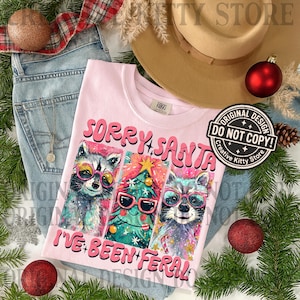 Puede incluir: Camiseta rosa claro con el texto "SORRY SANTA I'VE BEEN FERAL" y un gr&aacute;fico de mapaches y un &aacute;rbol de Navidad con gafas de sol. La camiseta est&aacute; sobre unos vaqueros azules.