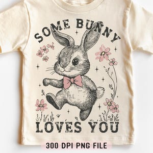 Peut inclure: Un t-shirt beige avec un motif de lapin portant un n&oelig;ud papillon rose. Le texte "Some Bunny Loves You" est imprim&eacute; sur le t-shirt dans une police de caract&egrave;res de style vintage. Le t-shirt est en 300 DPI.